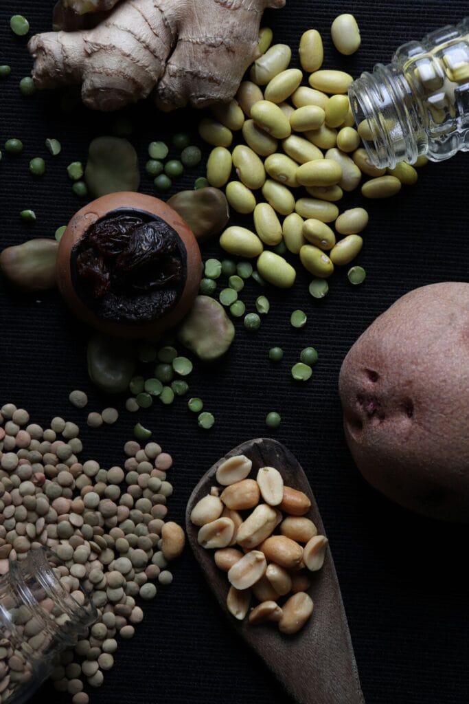 Peulvruchten van bovenaf op een donkere ondergrond: gele bonen en linzen die uit omgevallen glazen potjes rollen, groene spliterwten, tuinbonen en pinda's op een houten lepel, met daarnaast een schaaltje gedroogd fruit, een stuk gemberwortel en een aardappel